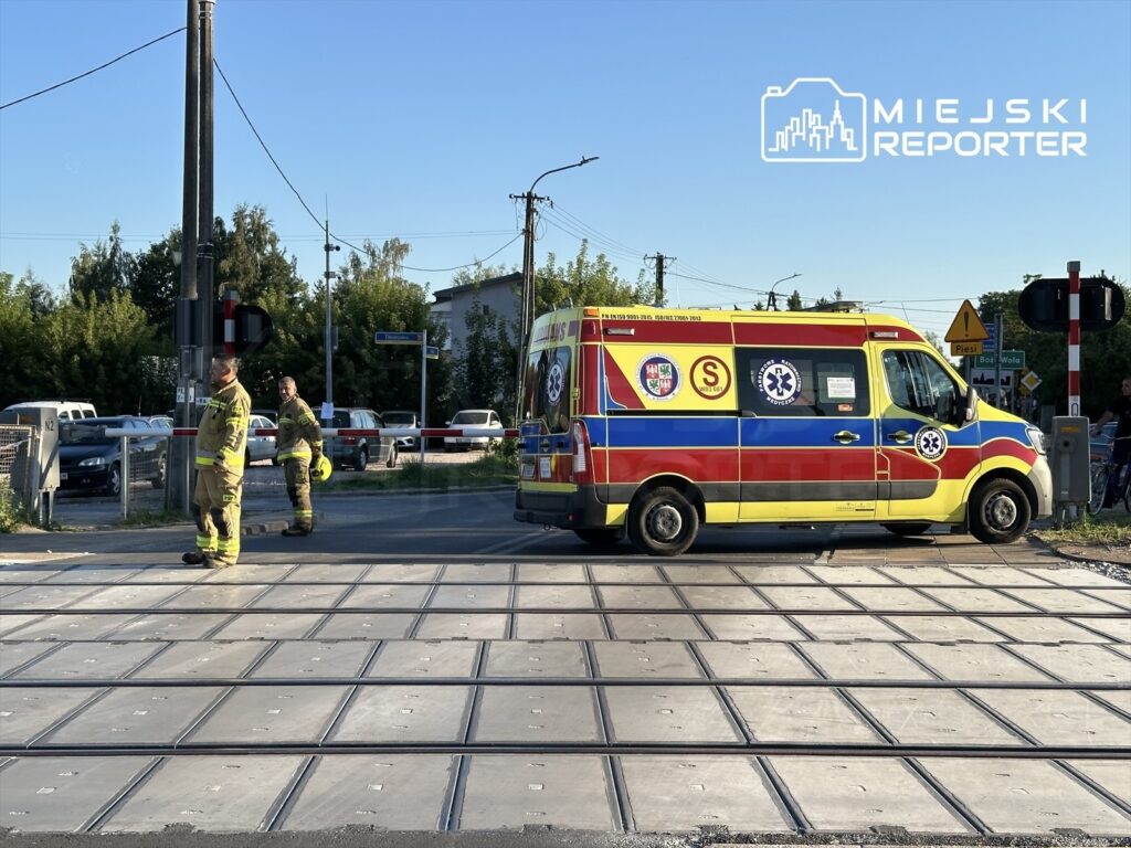 Zespół ratunkowy w specjalnych strojach stoi obok karetki przy przejeździe kolejowym.
