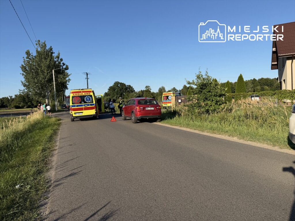 Na drodze stoi ambulans, obok grupka osób oraz dwa samochody, w tym czerwony pojazd z włączonymi światłami awaryjnymi.