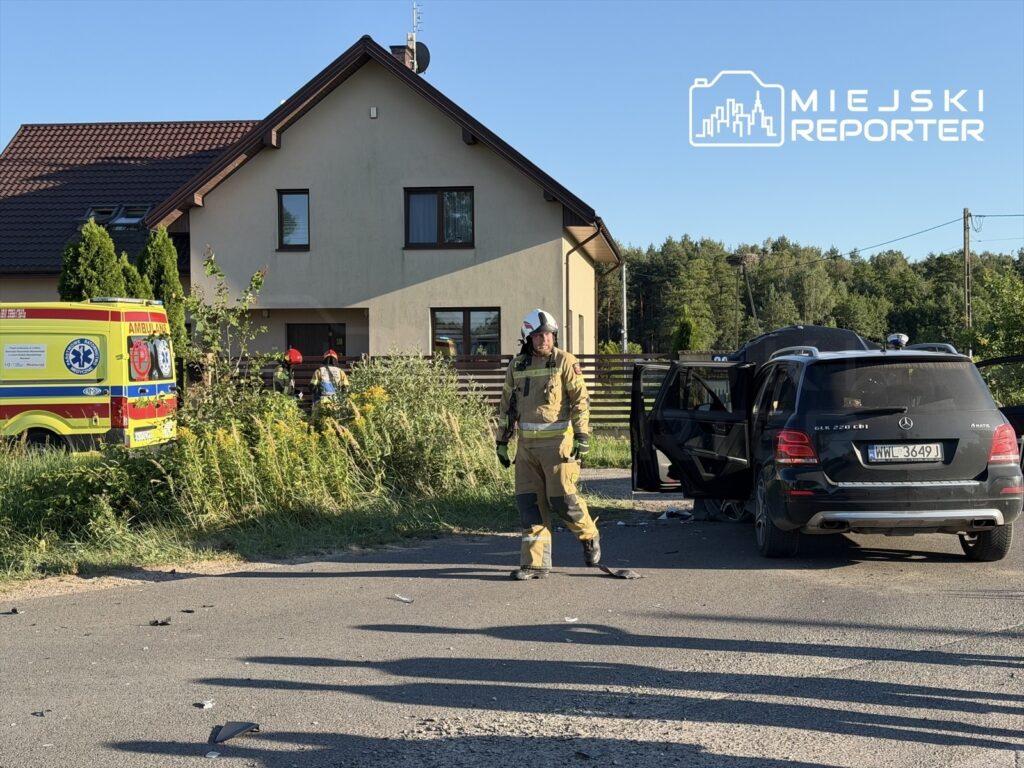 Strażak w żółtym mundurze stoi obok dachującego Mercedesa przy przydrożnym rowie, w tle karetka pogotowia.