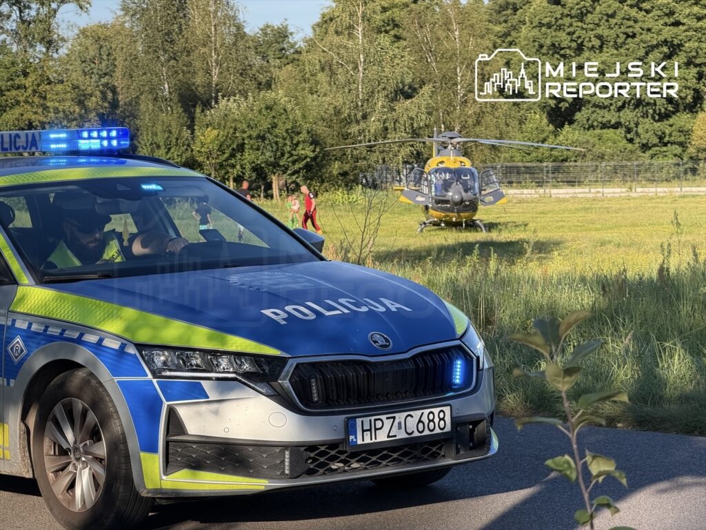 Policyjny radiowóz z włączonymi sygnałami świetlnymi stoi na poboczu drogi, a w tle lądował śmigłowiec ratunkowy.