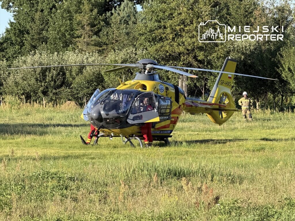 Lądowanie śmigłowca ratunkowego na zielonym terenie, w tle widoczni strażacy w mundurach.
