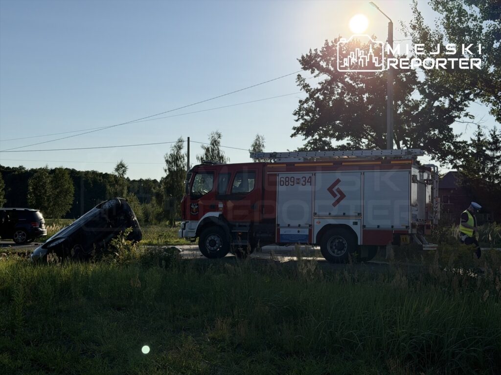 Czarny samochód osobowy dachujący w rowie przy drodze, obok strażacki wóz ratunkowy oraz funkcjonariusz w kamizelce odblaskowej.