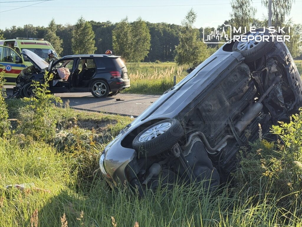 Dachowane auto osobowe leży w przydrożnym rowie, obok stoi rozbite SUV z otwartą maską, a w tle widać ratowników.