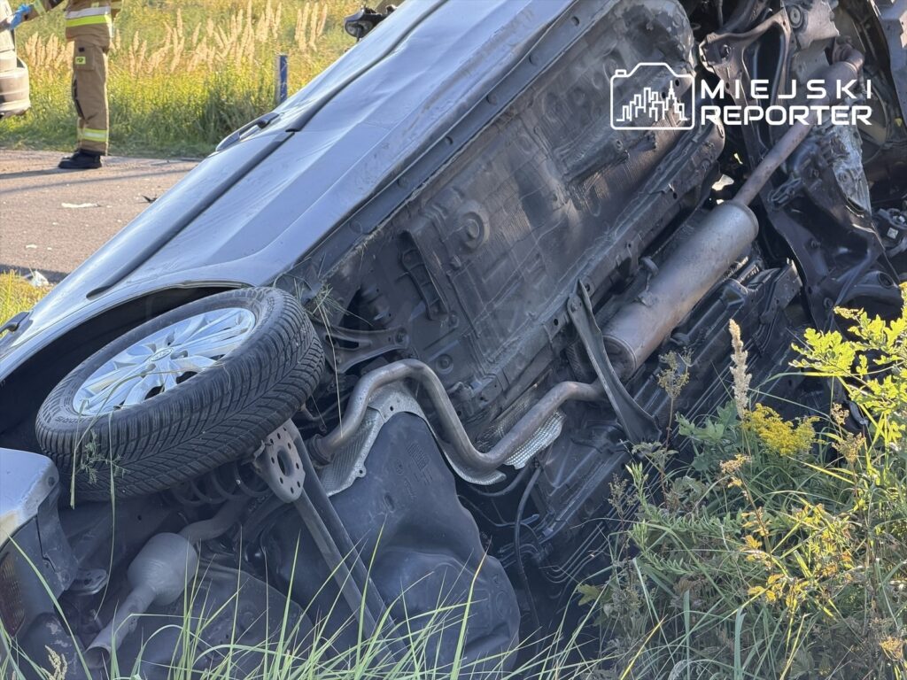 Czarne auto dachujące w przydrożnym rowie, obok strażak w mundurze, trawa i krzewy w tle.