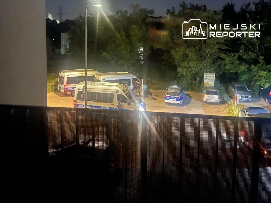 Grupa radiowozów policyjnych oraz samochodów służbowych zaparkowanych na oświetlonym placu nocą.