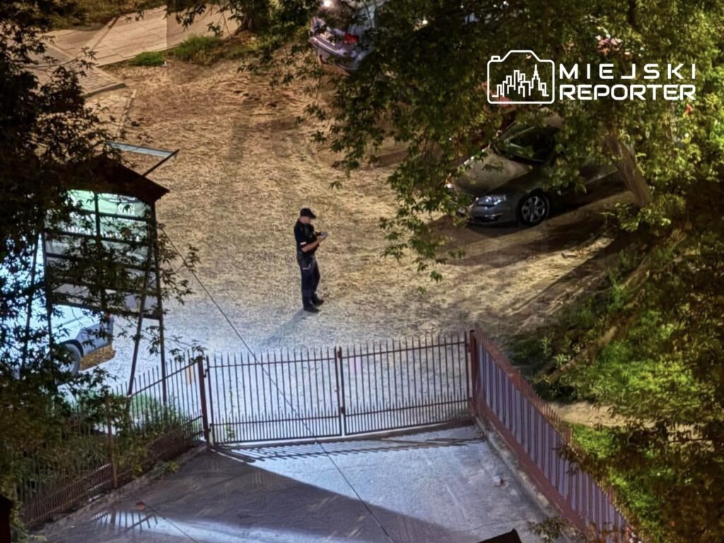 Policjant w mundurze przegląda notatki na opustoszałym parkingu, obok zamkniętej bramy.