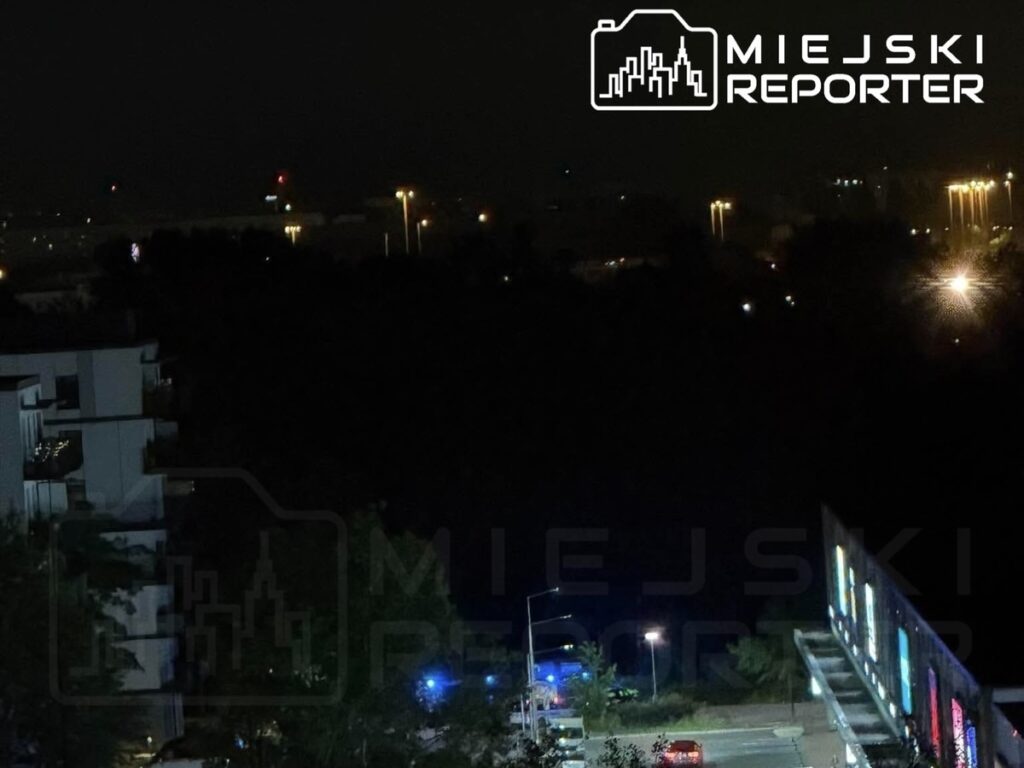 Nocny widok na miasto z oświetlonymi ulicami, w tle widać pojazdy służb ratunkowych oraz nowoczesny budynek.