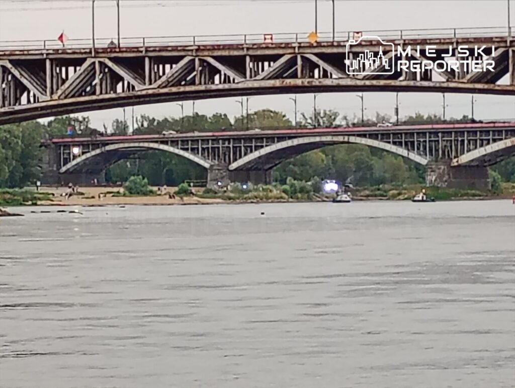 W tle mostu, łodzie ratunkowe przeszukują nurt rzeki, w pobliżu plaży z ludźmi i zielenią.