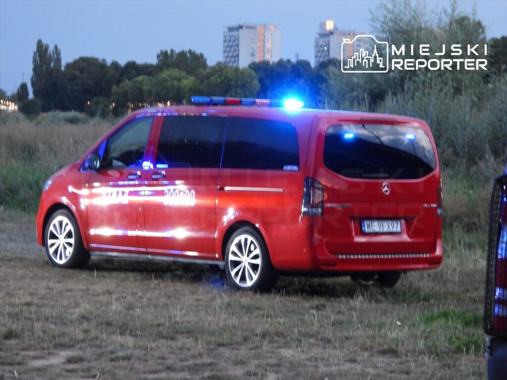 Czerwony samochód służb ratunkowych z włączonymi sygnałami świetlnymi stoi na trawie w pobliżu rzeki.