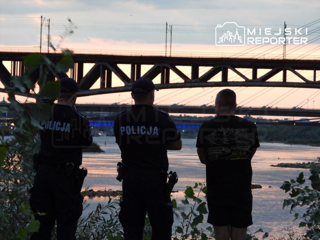 Trzech policjantów w mundurach obserwuje rzekę z brzegów, w tle widoczny most i zachodzące słońce.