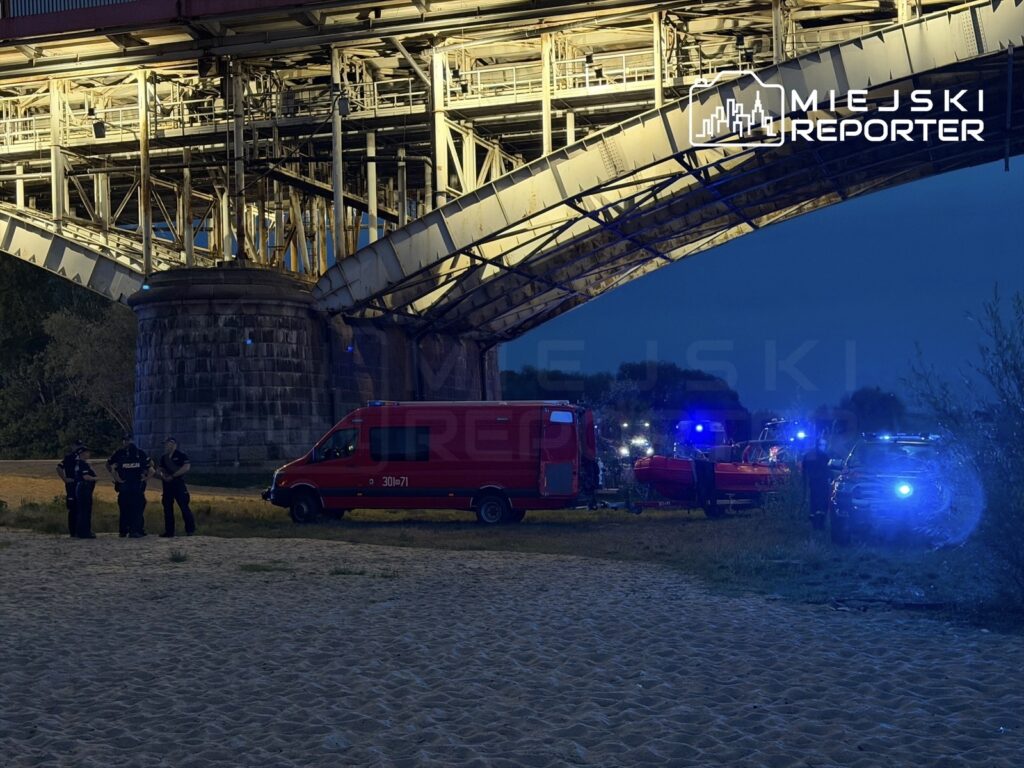 Grupa policjantów rozmawia obok wozu strażackiego i łodzi ratunkowej pod mostem, w nocy.