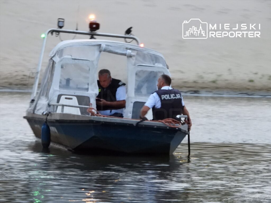 Dwóch policjantów na łodzi patrolowej przeszukuje wodę w poszukiwaniu zaginionego mężczyzny. W tle widoczny piasek i brzeg rzeki.