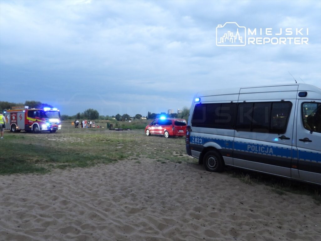 Na plaży w pobliżu rzeki stoją pojazdy służb ratunkowych, a w oddali grupka ludzi obserwuje sytuację.