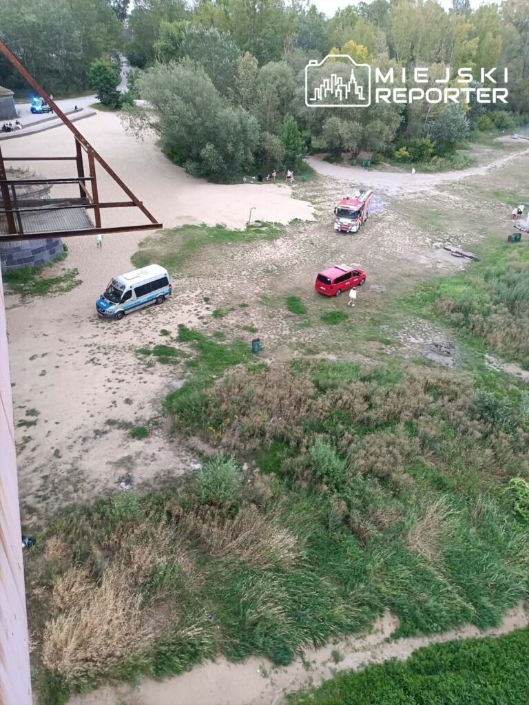 Na piaszczystym terenie wzdłuż rzeki stoją dwa samochody, w tym jeden strażacki, a w tle widać zieleń drzew.