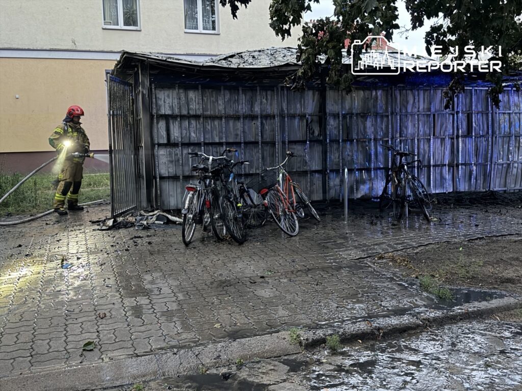 Strażak w hełmie gasi płonącą altanę obok rowerów na mokrej, wyłożonej kostką brukową ulicy.
