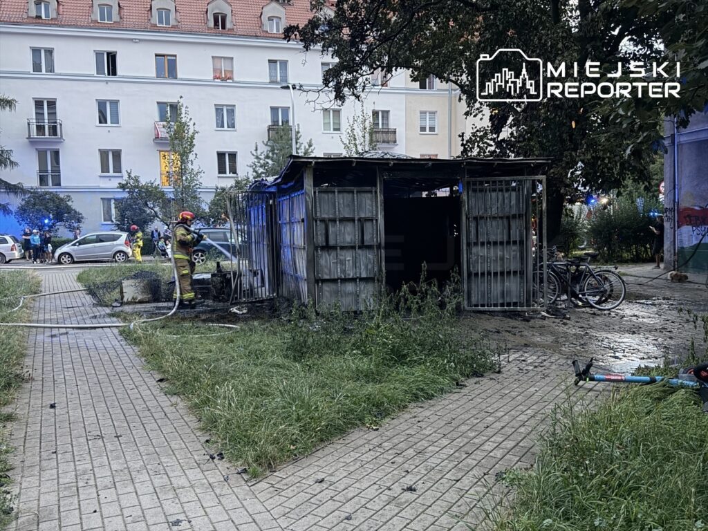 Strażak w hełmie gasi pożar altany, w tle widoczne zniszczone rowery i zespół ratunkowy na Mokotowie.