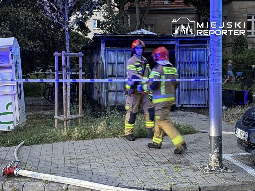 Dwaj strażacy w mundurach z hełmami podchodzą do altany, zabezpieczając miejsce po pożarze. W tle widoczne rowery.