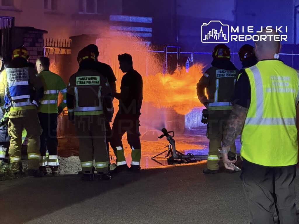 Grupa strażaków w mundurach, z wężami pożarniczymi, walczy z ogniem w nocy przy ogrodzeniu budynku.