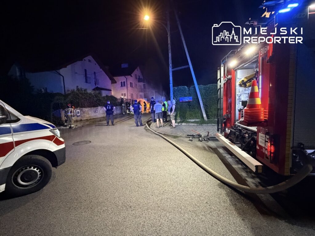 Grupa ratowników w kamizelkach odblaskowych pracuje w nocy przy wozie strażackim na oświetlonej ulicy.