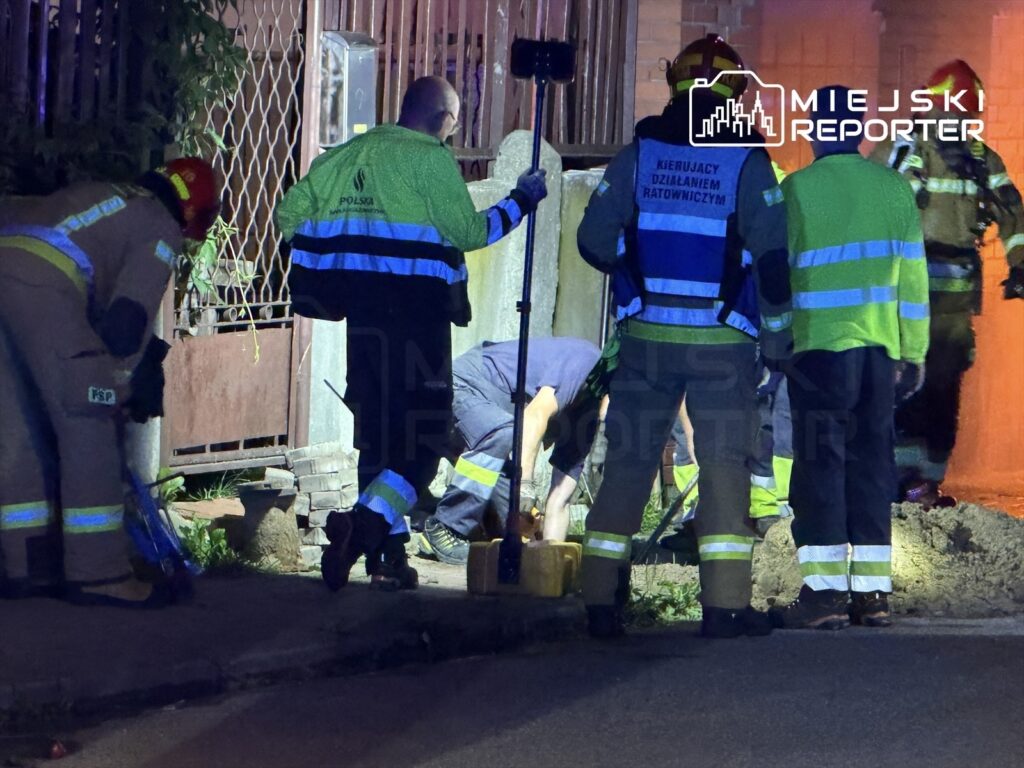 Grupa strażaków w mundurach, pracująca przy otwartym wykopie na ulicy, w nocy, z użyciem narzędzi ratunkowych.