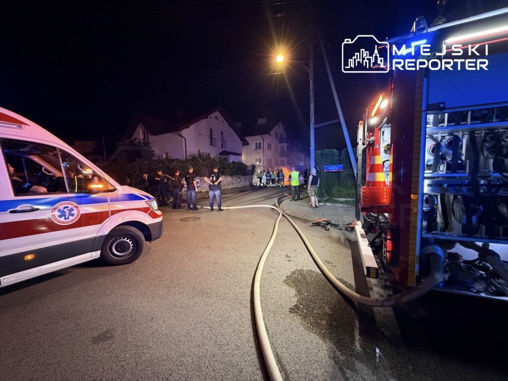 Zespół ratunkowy i strażacy pracują na miejscu zdarzenia w nocy, otoczeni przez pojazdy służbowe i gapiów.