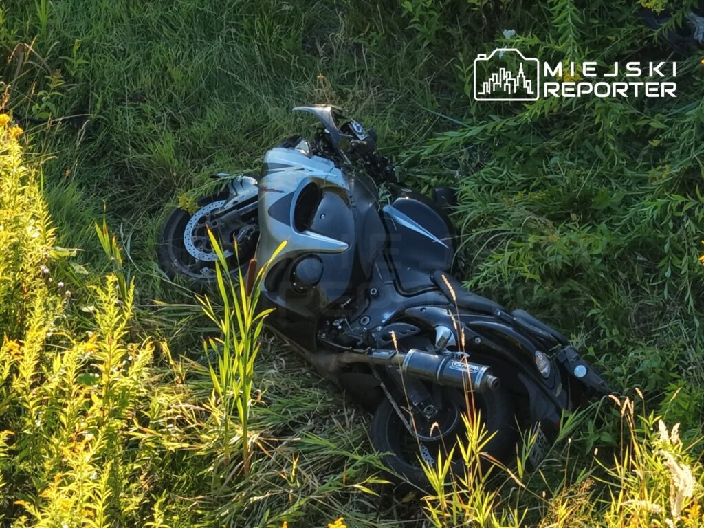 Motocykl leży w przydrożnym rowie, otoczony wysoką trawą i roślinnością.