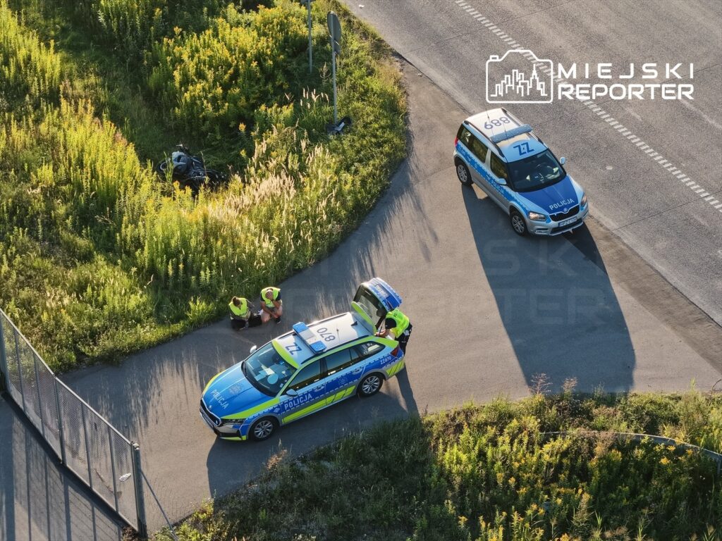 Policjanci w odblaskowych kamizelkach prowadzą działania na poboczu drogi, obok przewróconego motocykla.