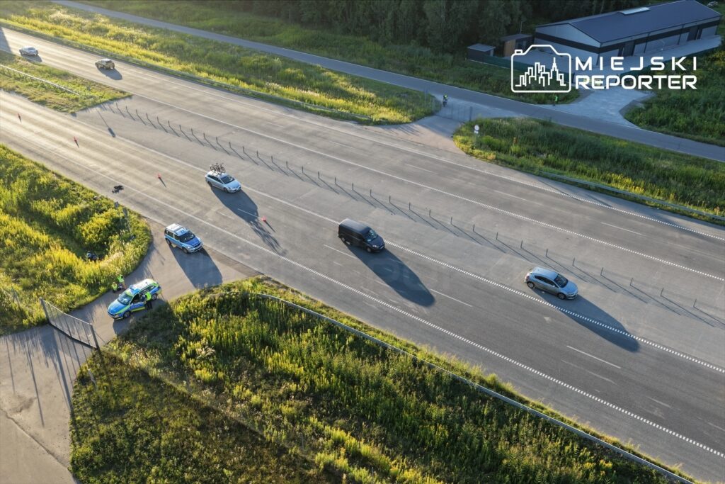 Na drodze widoczne są pojazdy, w tym radiowóz, w otoczeniu zieleni i pasów wyznaczających ruch.
