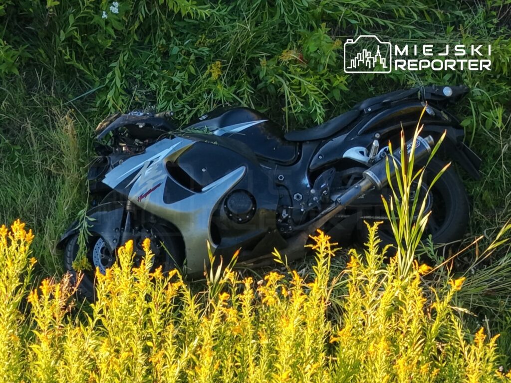 Motocykl leżący w przydrożnym rowie, częściowo zasłonięty przez wysoką trawę i kwiaty.