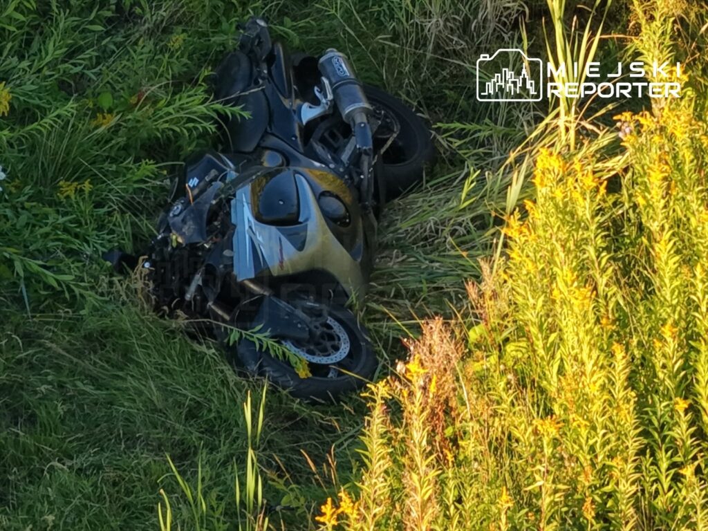 Zniszczony motocykl leży w wysokiej trawie przy drodze, otoczony dzikimi kwiatami.