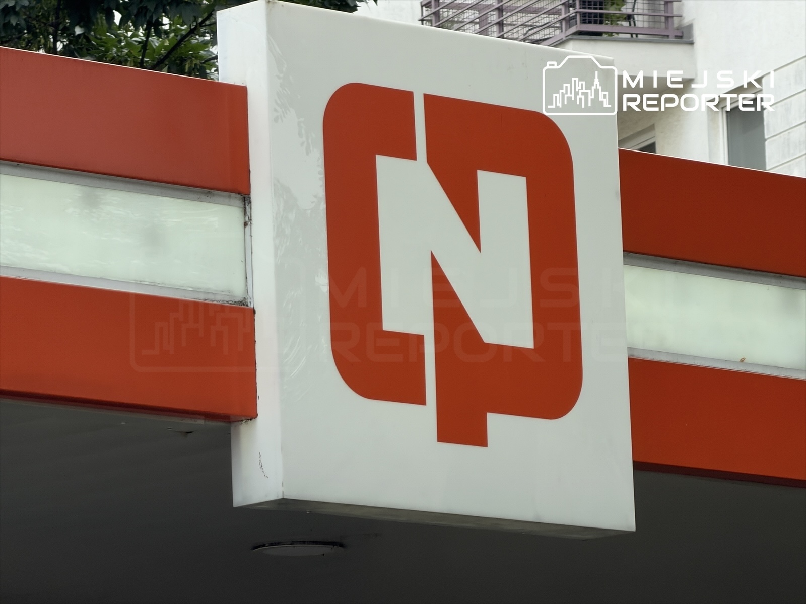 Logo z literami "N" i "P" na białym tle, umieszczone na czerwonym tle, w miejskim otoczeniu.