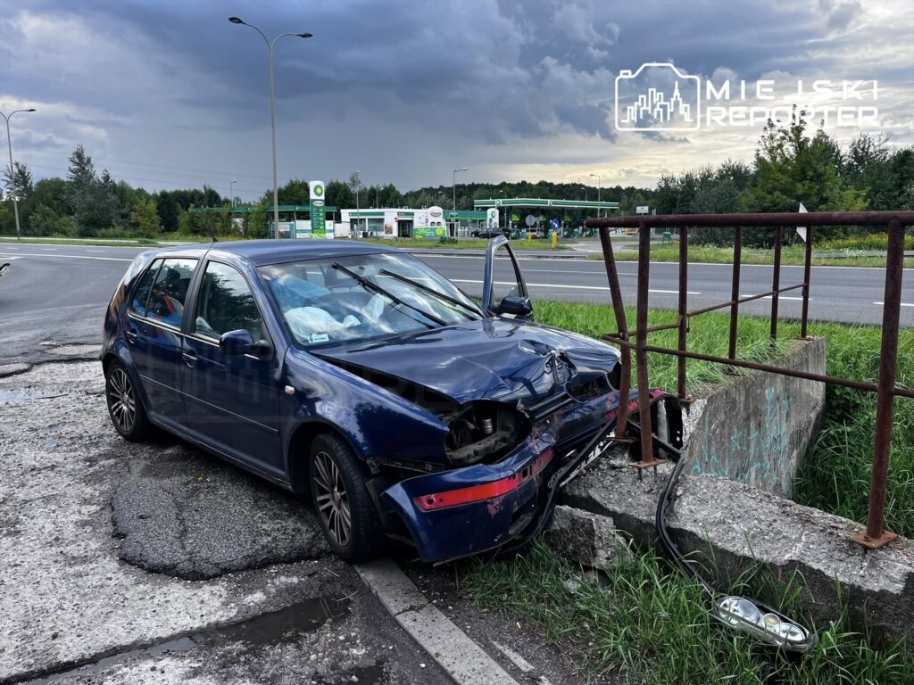 Zniszczony niebieski Volkswagen leży na poboczu drogi, uderzony w betonowy przepust, obok widoczna stacja benzynowa.