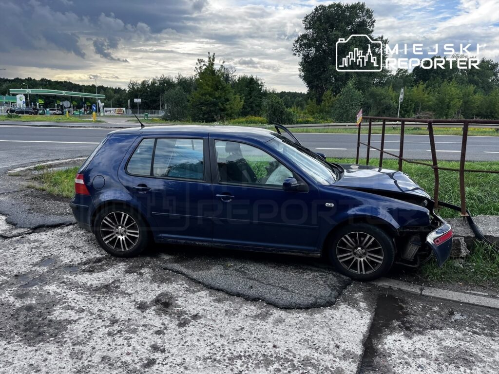 Niebieski Volkswagen leży na poboczu drogi, z uszkodzoną przednią częścią, obok betonowego przepustu i ogrodzenia.