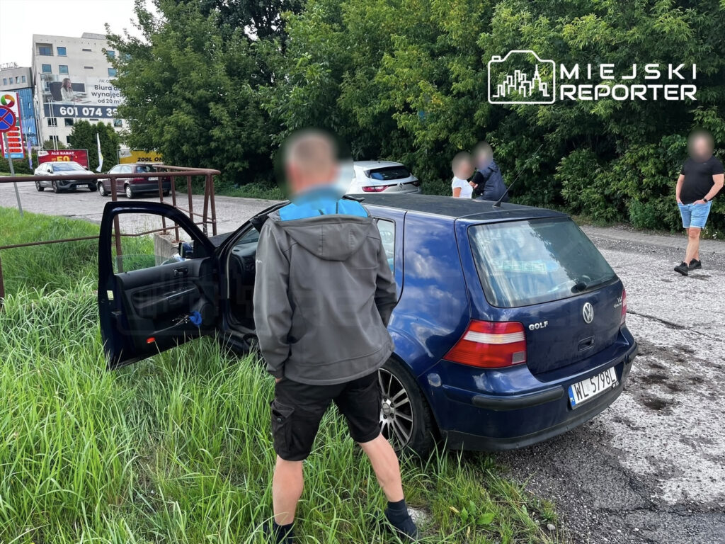 Mężczyzna w szarym płaszczu stoi obok niebieskiego Volkswagena Golfa, którego drzwi są otwarte, na trawiastej poboczu drogi.
