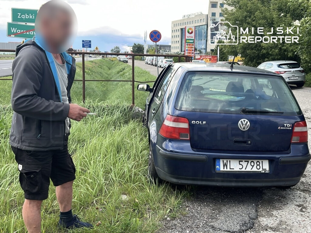 Mężczyzna w szarej bluzie stoi obok uszkodzonego samochodu Volkswagen Golf, zaparkowanego na poboczu drogi.