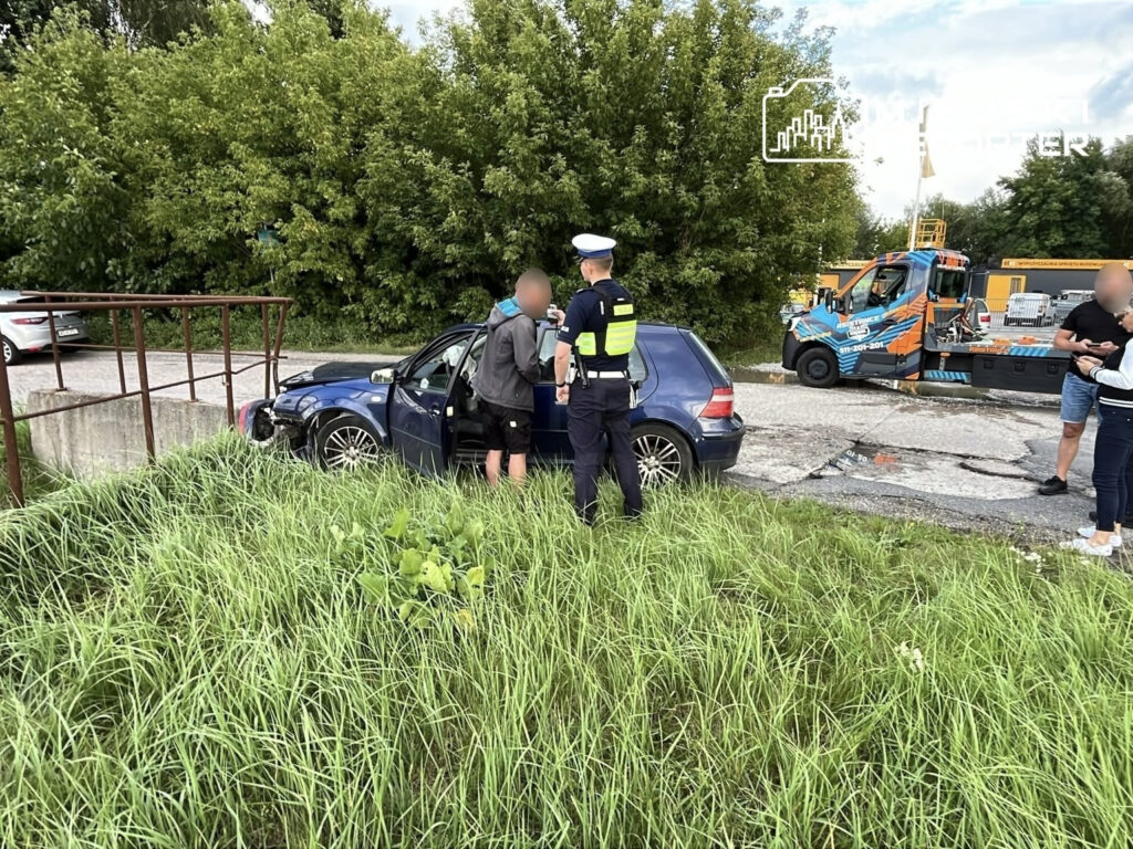 Kierowca w szarej bluzie rozmawia z policjantem obok uszkodzonego niebieskiego Volkswagena w zaroślach.