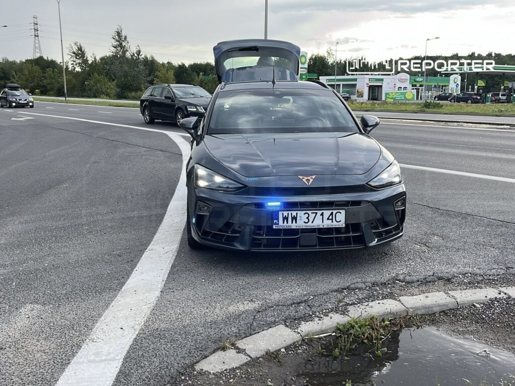 Czarny samochód osobowy z włączonymi sygnałami świetlnymi stoi na poboczu drogi, obok stacji paliw.