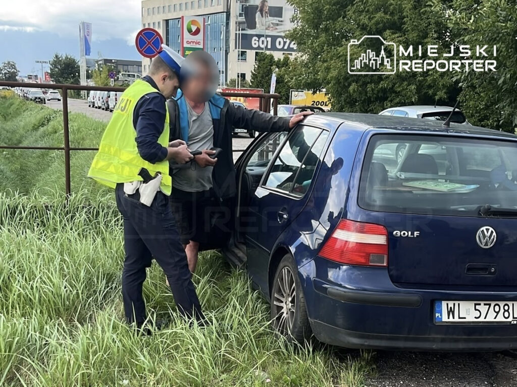 Policjant w kamizelce odblaskowej rozmawia z mężczyzną przy niebieskim Volkswagenie Golfie zaparkowanym na poboczu.