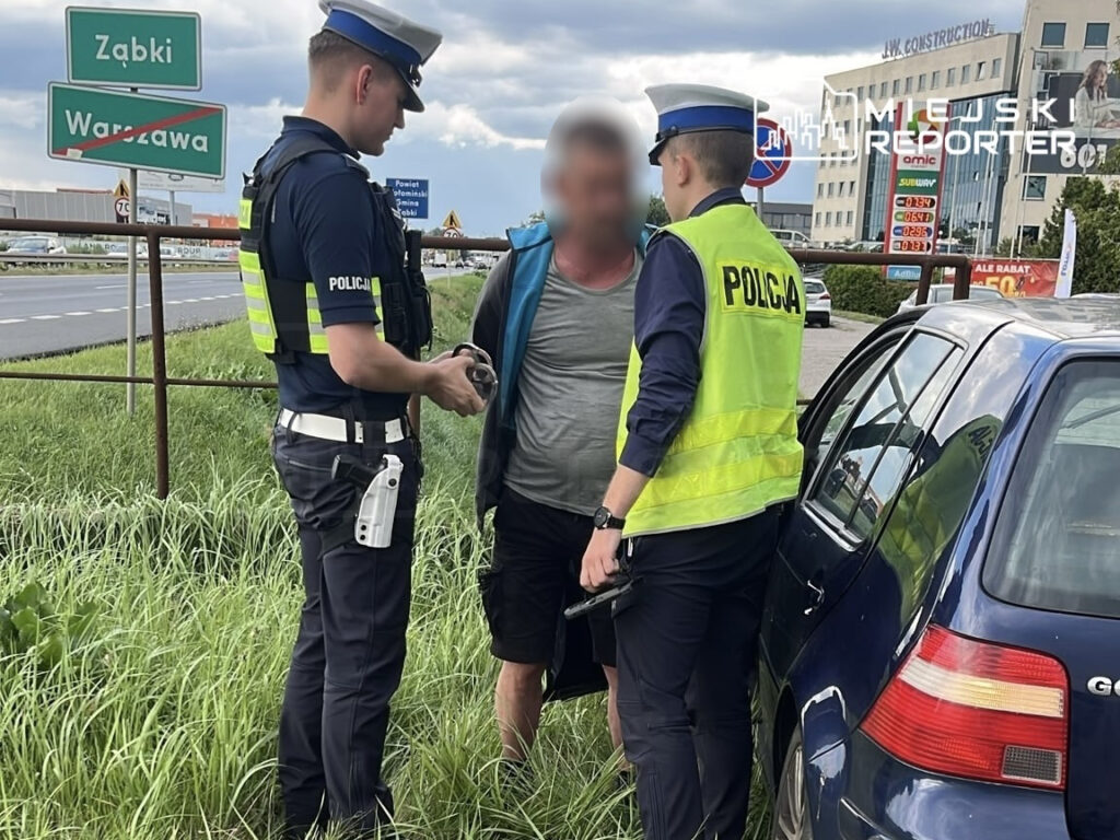 Mężczyzna w szarej koszulce jest zatrzymywany przez dwóch policjantów na poboczu drogi, obok niebieskiego samochodu.