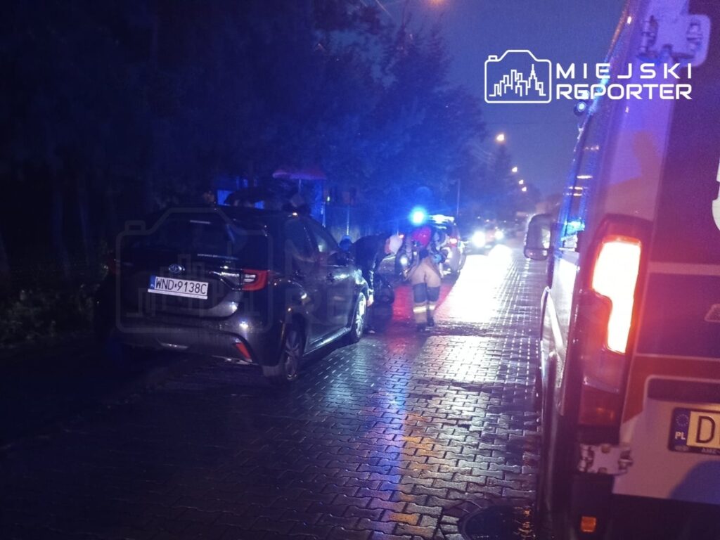 Na mokrej nawierzchni ulicy, ratownicy medyczni udzielają pomocy poszkodowanemu obok dwóch uszkodzonych pojazdów.