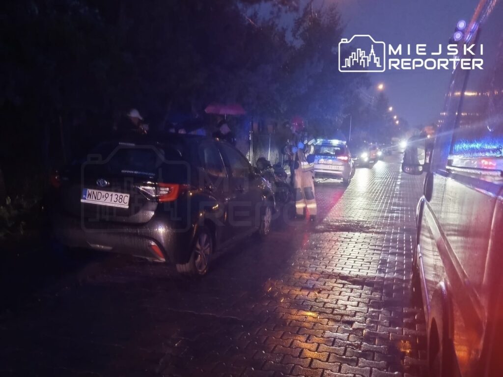 Grupa ratowników w odblaskowych strojach bada miejsce zdarzenia na deszczowej ulicy, obok uszkodzonych pojazdów.