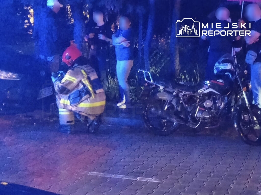 Strażak w mundurze klęczy obok uszkodzonego samochodu, podczas gdy grupa osób obserwuje sytuację w nocy.