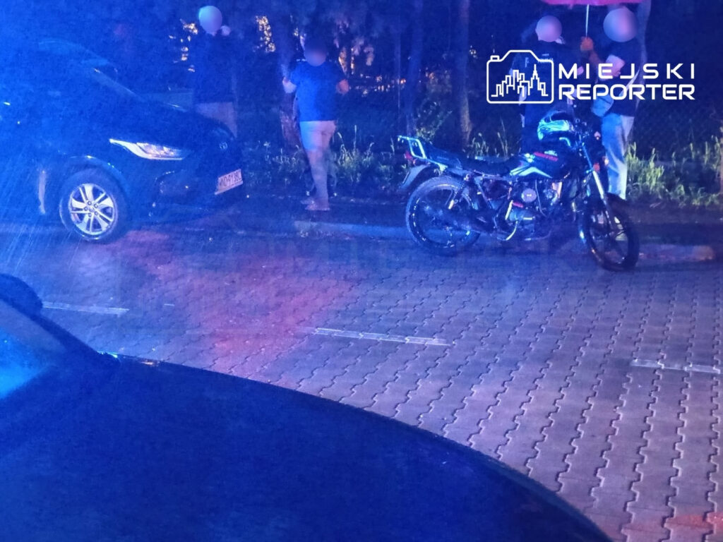 Grupa osób stoi obok czarnego samochodu, a obok nich zaparkowany motorower. Scena oświetlona niebieskimi światłami.