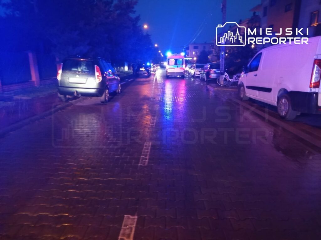 Na oświetlonej ulicy zaparkowane są samochody, a w tle widać policyjne radiowozy z włączonymi sygnałami świetlnymi.