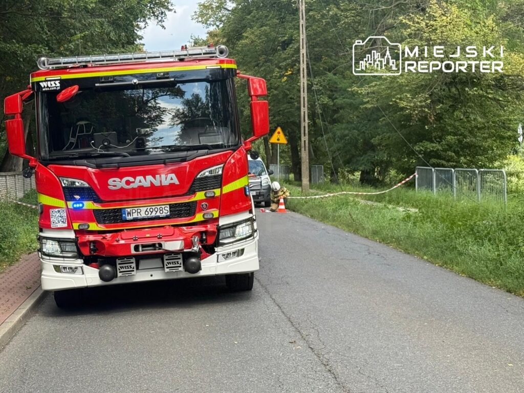 Czerwony wóz strażacki marki Scania zatrzymany na drodze, obok oznakowanie ostrzegawcze i taśma zabezpieczająca.