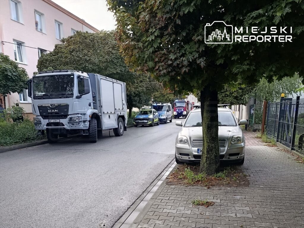 Na ulicy stoją pojazdy służb ratunkowych, w tym duża ciężarówka oraz radiowóz, otoczone drzewami i budynkami mieszkalnymi.