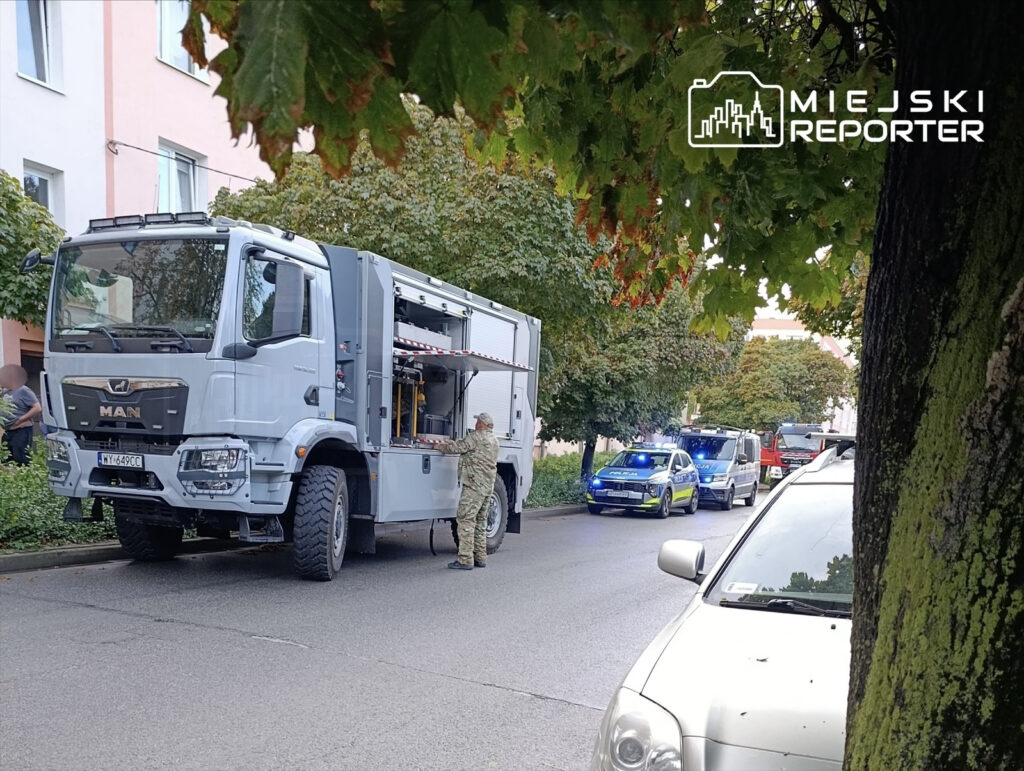 Kontrterroryści w mundurach sprawdzają sprzęt w specjalistycznym pojeździe na ulicy otoczonej drzewami.