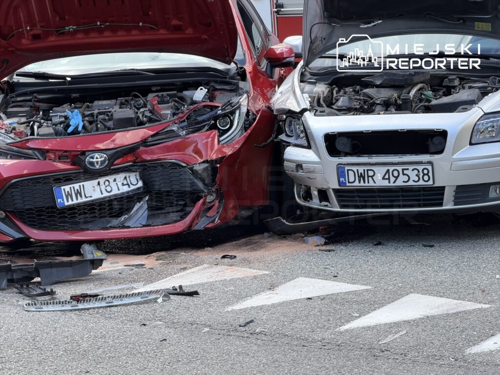 Dwa uszkodzone samochody osobowe zderzone na drodze, jeden czerwony, drugi srebrny, z widocznymi uszkodzeniami przodu.