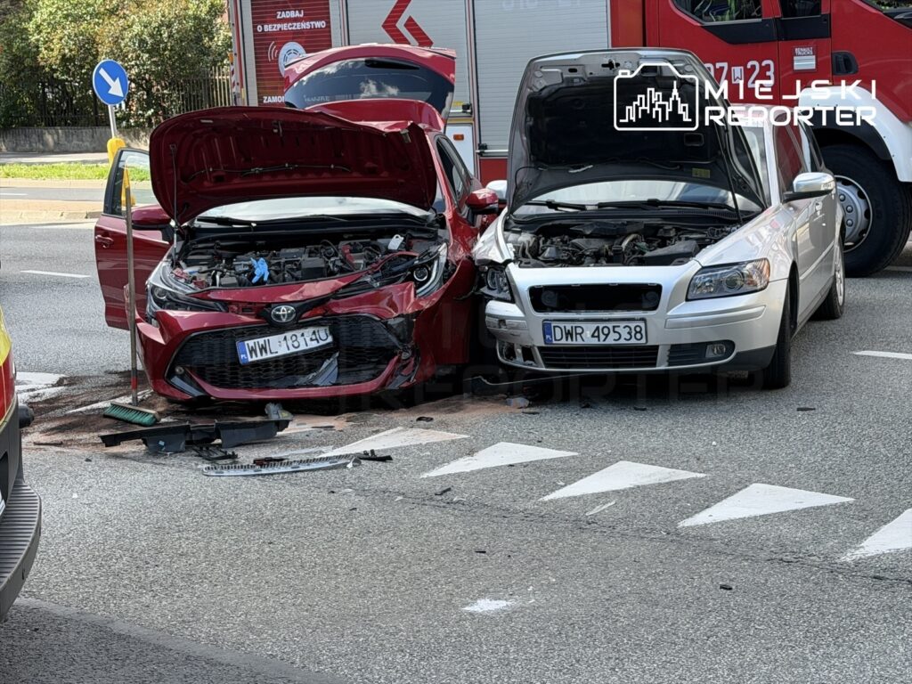 Dwa uszkodzone samochody osobowe z otwartymi maskami na ruchliwej ulicy, obok znak drogowy i sprzęt do sprzątania.