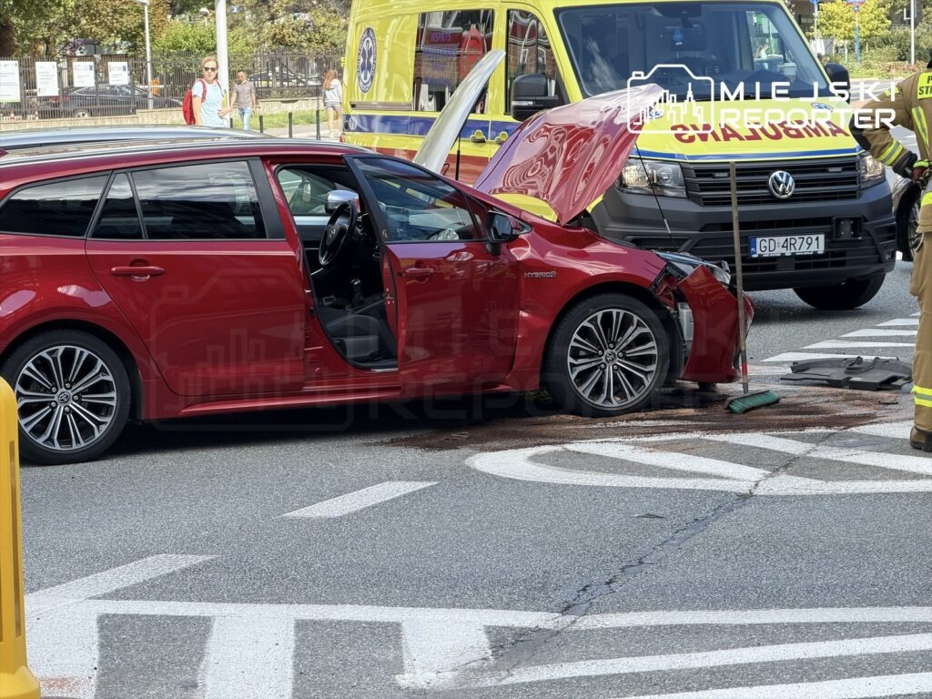Czerwony samochód osobowy z uszkodzoną przednią częścią stoi na jezdni, obok pracują strażacy i karetka.